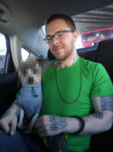 A photo of Ryan Michael Irwin, taken during or before March 2020. Taken from what appears to be the driver's seat, Irwin sits in the back passenger side of a car. On his lap is a Jack Russel terrier, wearing a gray hoodie with green hood pulls; the dog's face has been intentionally redacted. Irwin imself wears blue jeans, an emerald green shirt, a black wristband loosely on his left arm, a chain of what appear to be gold plastic Mardi Gras beads, and two-tone horn-rimmed corrective lenses with rectangular frames. He has a short blond beard and a buzzcut, his face wearing the ghost of a smirk. Several of Irwin's tattoos are visible, including the heart-and-triquetra on the right side of his neck and the star peeking out of his shirt collar. On the back of his left hand is what appears to be a flower design with the Traditional Chinese characters "熱情" on it; this roughly translates to "passion" or "enthusiasm" by way of fervor or zeal. His arms are adorned with several other tattoos, though they're difficult to make out.