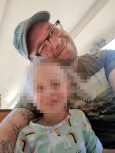 A selfie of Ryan Michael Irwin taken from below, framing him from the hips up from a sitting position, during 2024. Here, Irwin is 37. In Irwin's arms appears to be a daughter, no older than three. Her face has been intentionally redacted. They're sitting in one of the cars of the Explorer Express Train at the Worcester EcoTarium, a wooden bench visible behind Irwin's right arm. Irwin wears an olive green graphic design tee, a green baseball cap with the word "Worcester" on it embroidered in script, a gold chain necklace, and his two-tone horn-rimmed glasses. His beard is short, and he is smiling broadly. A tattoo on Irwin's right bicep appears to depict the bust of a vaguely feline woman, her face and cleavage on the same broad bodily plane and surrounded with fur, just next and under his daughter's face.