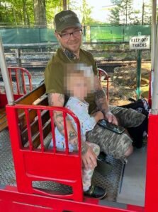 A photo of Ryan Michael Irwin and his daughter as they ride the Worcester EcoTraium's Explorer Express Train, during 2024. They both face the camera, which is outside the train. The daughter's face has been intentionally redacted. Irwin wears an olive green graphic design tee, a green baseball cap with the word "Worcester" on it embroidered in script, a gold chain necklace, his two-tone horn-rimmed glasses, olive green camo shorts, and what appear to be black Crocs. Irwin smiles faintly toward the camera, his smartphone in his lap. Several of his tattoos are visible along his arms and neck. The combination heart-and-triquetra on the right side of his neck has been printed over with a valknut. On the back of his right hand is an angrily grinning skull wreathed in flames. Above that on his right forearm is a puzzle piece, depicting what appears to be a cloudy sky. Toward his right elbow is a large dove. On the inside of his left elbow is what appears to be a highly stylized face surrounded by large white stars, a grid pattern being chewed between its jaws.