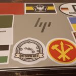 The first photo of Colwell's laptop, displaying the backside of the monitor. It includes decals of the New England Independence flag, the Vermont Republic flag, the Appeal to Heaven flag, the Korean Workers' Party insignia, the Palestinian flag, an emblem of an ouroboros encircling the Big Dipper constellation with text asking "Qvis contra nos?" ("who can be against us?"), the Ukraine flag, the flag of the fictional Russian warlord Black League state, and what appears to be an Azov Battalion patch minus the text and the vertical bar of the central wolfsangel symbol. There is also a generic Deutschland decal featuring two different versions of the German flag.