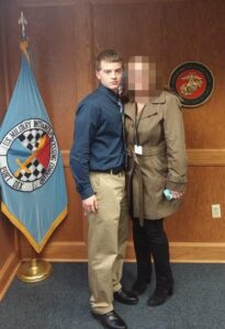 A photo of Brandon Brooks circa 2017, standing with his mother in what is apparently Fort Dix. To his right is a Fort Dix flag. To his left is his mother, her face redacted. To her  left is a wall emblem for the Department of the Navy United States Marine Corps.