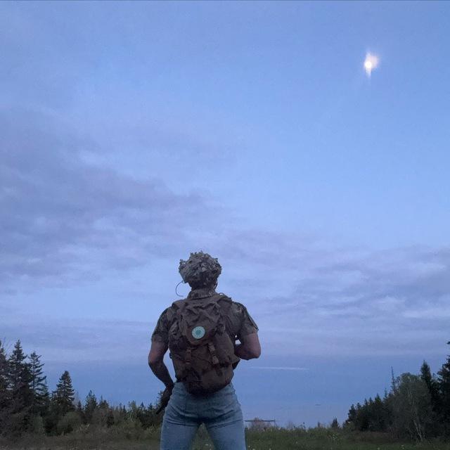 A photo of who is apparently Brandon Brooks, dressed in a tactical vest and holding what appears to be a carbine rifle. He is staring off into the sky, his back to the camera. A patch on his tactical backpack appears to be a green-and-white sonnenrad.