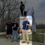 A photo from the New England National Party's website gallery, taken during the 250th Anniversary of Concord MA during April 2025. Three white men stand in a row in front of the Minute Man statue in the historical park of the same name. The man on the left is an unknown. In the center is PINE member alias "Adam Smith". On the right is Bobby Garcia, in khaki pants, work boots, and navy blue tanktop, bearing a flag of the NENP's stag logo.