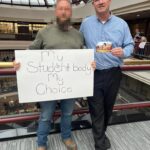A photo taken from the New England National Party's website gallery, taken during a legislative session at Hartford CT during May 2025. Bobby Garcia, wearing a light blue dress shirt and black dress pants, stands next to a homeschooling advocate. The advocate holds a sign that says "My Student Body My Choice". Garcia holds an NENP propaganda flyer.