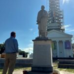 A photo from the New England National Party's website gallery, taken outside of the Enfield Congregational Church in Enfield CT during June 2025. Bobby Garcia, with his back to the camera, stands facing the statue of Thomas Abbey in front of the church. Visible in the background, the church's steeple is wreathed in scaffolding for a repair project; from that scaffolding hangs a rainbow Pride flag.