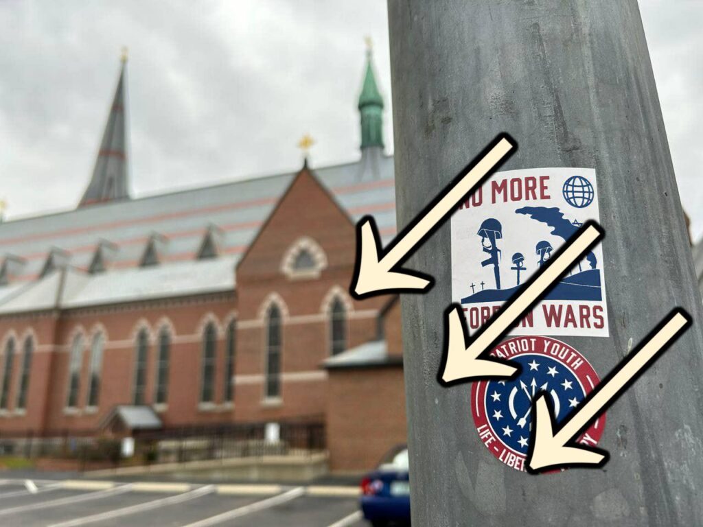 A photo taken near St. Joseph's Cathedral in Hooksett NH sometime around 2022 or 2023, with the cathedral visible in the background. In the foreground is a street lamp pole defaced with two white nationalist stickers. One is a "Roblox Youth" sticker, and the other is a Patriot Front "No More Foreign Wars" sticker. The stickers have a set of three arrows, pointing downward and to the left, superimposed over them.