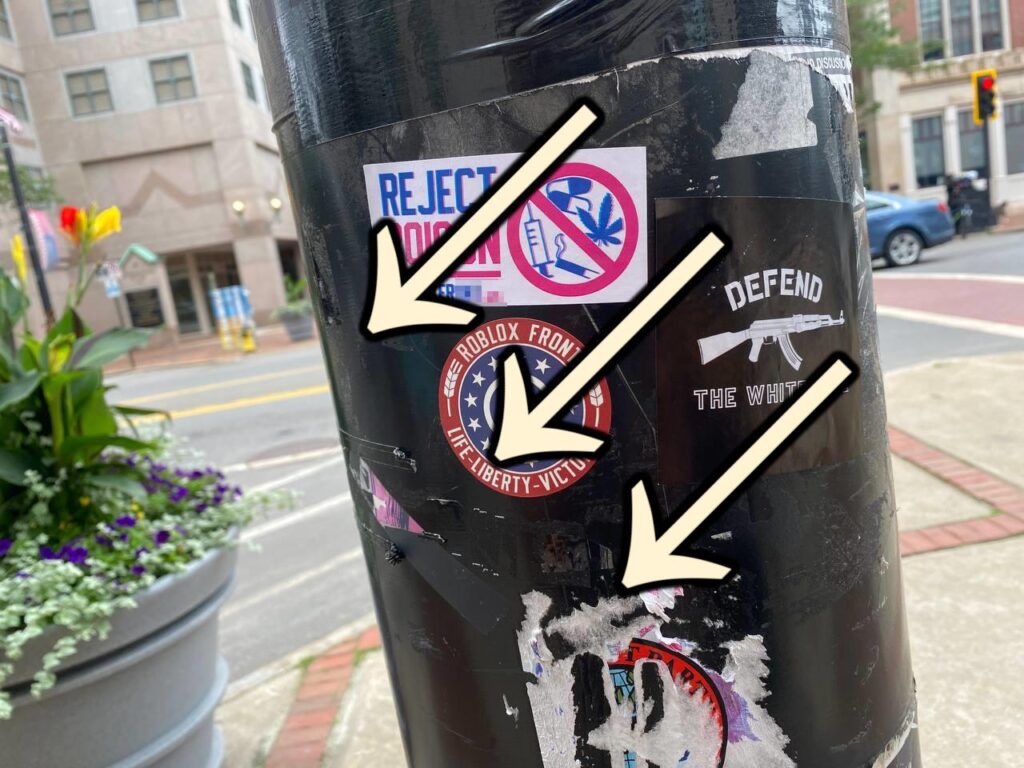 A photo taken at Manchester City Hall Plaza sometime around 2022 or 2023, with the 900 City Hall Plaza building visible in the background. In the foreground is a street lamp pole defaced with three white nationalist stickers. One is a "Roblox Front" sticker, another is a Patriot Front "Reject Poison" sticker, and the third juxtaposes an image of an AK-47 with the text "Defend the White Race." The stickers have a set of three arrows, pointing downward and to the left, superimposed over them.