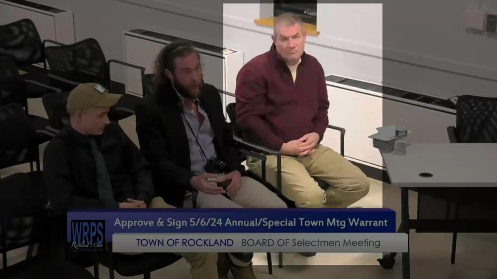 A screenshot of a Rockland MA town meeting during May 2024. Among the largely unoccupied seating sit three white men from NSC-131's PINE group, there to bird-dog the city council about the presence of immigrants and refugees. Sitting to the right of the other two is Bobby Garcia, wearing khaki pants and a burgundy overshell. Sitting to his right are PINE members alias "James Ambrose" and alias "Adam Smith".