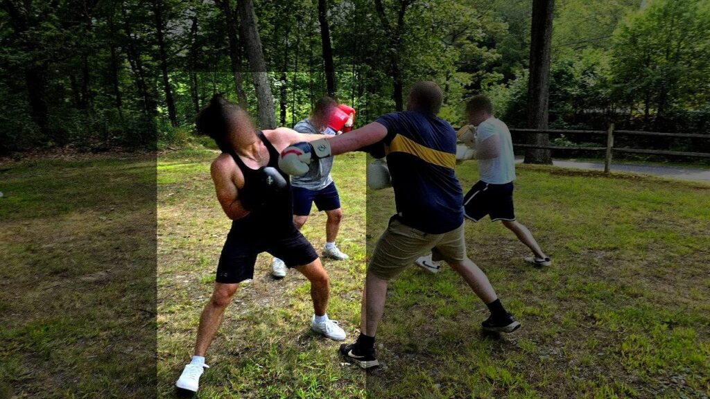 A photo taken in a park in Worcester MA, during August 2025, and in full color, a white man in what a black tank-top, black UnderArmour shorts, white sneakers, and black boxing gloves. He is in a wild half-squatting stance as he tries not to get punched in the face by the man to his right, likely to be Patriot Front member Joseph Behling of Manchester NH. His face has been blurred, his luxuriously full and dark hair visible. As he moves to not take a punch, his hair tossed in the air, going from luxurious to comical.