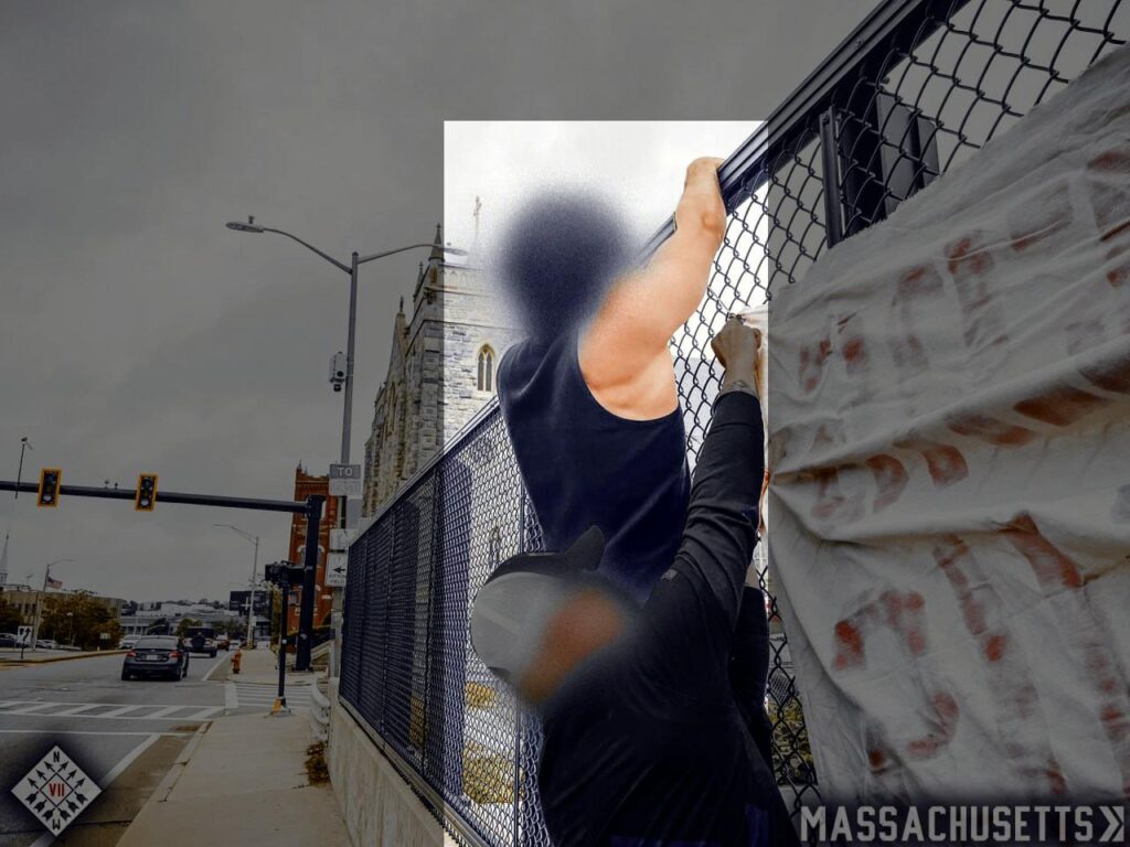 A photo of two white men tying a banner to a chain-link fence on an overpass in Worcester MA during June 2025. One of them, highlighted but largely occluded from view by the man in front of him, wears a black tanktop. He has dark hair.