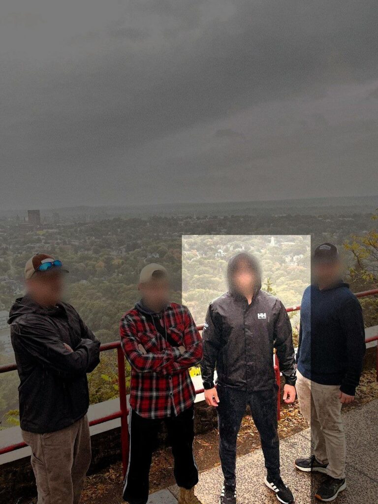 A photo of four white men posing on the path to the Soldiers & Sailors Monument in New Haven CT, a view of the city below behind them. One, highlighted, wears a black Helly Hansen raincoat, black training pants, and black Adidas trainers. His face is blurred. Standing with him are what appear to be three other members of the New England Active Club, including Mark Hayden of Spencer MA.