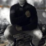 A photo of a Patriot Front member taken sometime during December 2022, somewhere in or around Manchester NH. A white man of average build squats in front of a concrete wall, holding his right fist to his heart. He wears what appears to be a Columbia Men's Ascender softshell jacket in navy blue, khaki pants, black minimalist Nike sneakers, a white gaiter covering the lower half of his face, and a tan cap with a Patriot Front flag patch on it. What is visible of the upper half of his face is blurred. He stands on what appears to be a brick sidewalk, in front of a concrete wall vandalized with graffiti.