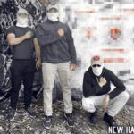 A photo of three Patriot Front members standing in front of a concrete wall in a wooded area somewhere in or around Manchester NH, taken during December 2022. Two men, who are apparently Patriot Front members Wyatt Trahan and Joseph Behling, stand next to a third man in a squatting position, who is apparently Jake Hayden. They are all white men, holding their right fists to their hearts, and concealing their faces with white gaiters and tan baseball caps with various patches on them. Trahan's patch blends in with his cap too well to be identifiable. Behling's patch is a black variant of the Vermont Republic flag. Hayden's is a Confederate navy jack flag. Their outfits are otherwise casual or sports wear. The wall behind them has been vandalized with graffiti, which has been graphically obscured.