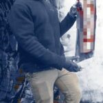 A photo of a Patriot Front member taken sometime during March 2023, somewhere in or around Manchester NH. A white man of average build stands facing the right, holding a graffiti stencil of some kind. He wears what appears to be a Columbia Men's Ascender softshell jacket in navy blue, khaki pants, black minimalist Nike sneakers, a white gaiter covering the lower half of his face, glasses, a tan cap with what appears to be an Appeal to Heaven flag patch on it, and protective rubber gloves smeared with white paint. He stands on what appears to be a brick sidewalk, in front of a concrete wall vandalized with graffiti.