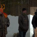 A still from a video taken during a Patriot Front hike somewhere in Maine during March 2023. A line of three apparent Patriot Front members stand in a snowy clearing near a treeline. They're all in various levels of winter clothing, some with hiking packs to their sides. One, a white man of average build highlighted in the still, wears khaki pants, a black Carhartt hooded winter jacket, and a black trapper hat with the earflaps down. While his face is blurred, there is still a resemblance between the Patriot Front member and the blithe grin occasionally photographed on the face of Joseph Behling.