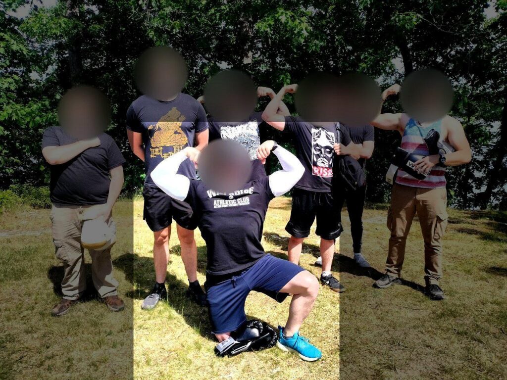 A photo of various Bay State Active Club members, all white men with blurred faces, posing somewhere in Worcester County MA, sometime during or before June 2024. One member, on one knee, flexes his biceps. He wears a black short-sleeve Will2Rise t-shirt, with a longsleeve white compression shirt underneath. His sneakers are a vivid light blue.