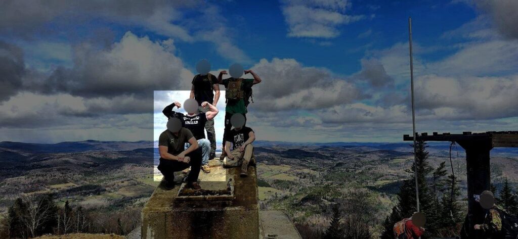 A photo of New England Active Club members, all white men with blurred faces, hiking somewhere in southern VT during or before April 2024. One member, on one knee, flexes his biceps. Wearing a black, short-sleeve Will2Rise t-shirt, a black undersleeve covers his right arm to the wrist.