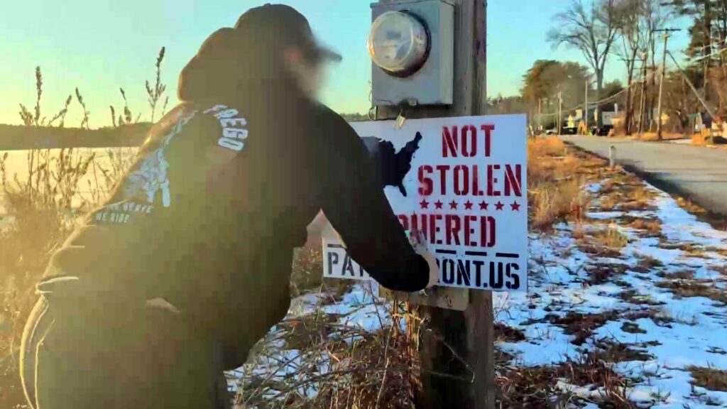 A photo of who is alleged to be Garrett Berna, somewhere in New England, during or before March 2025. It is visibly winter, with snow on dying grass, and barren trees in the background. The man alleged to be Berna staples a "Not Stolen, Conquered" Patriot Front sign to a utility pole.