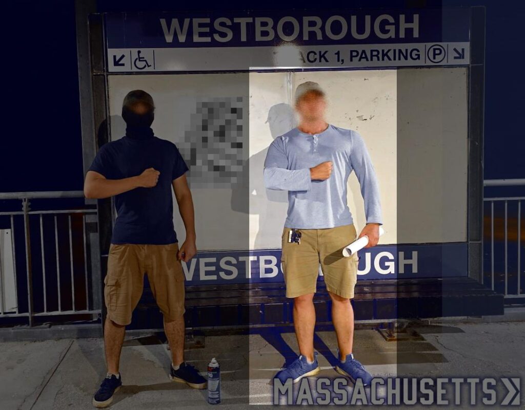 A photo of two members of Patriot Front at the Westborough MA commuter rail station, during or before September 2024. One member wears black khaki cargo shorts, black athletic sneakers, a black short-sleeve t-shirt, and a black balaclava. The other, alleged to be Garrett Berna, wears a light blue long-sleeve Henley t-shirt, khaki shorts, a tan baseball cap, and blue sneakers. Behind them is a Patriot Front poster (heavily pixelized and defaced), pasted to one of the rail station signs.