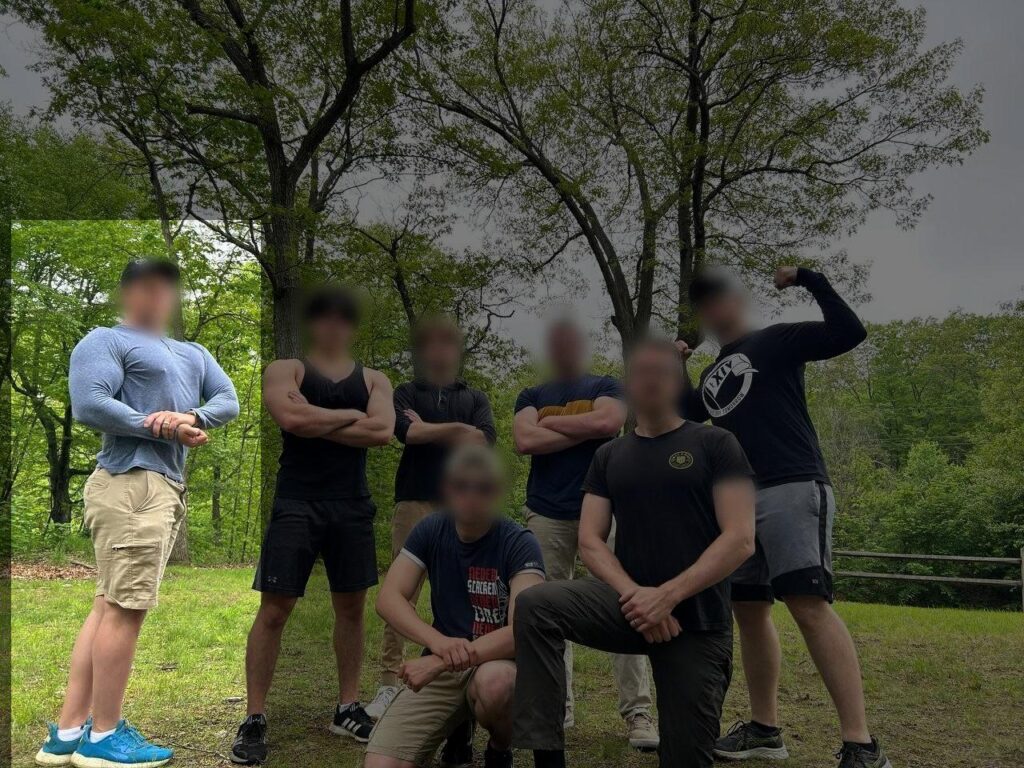 A photo of various Bay State Active Club members, all white men with blurred faces, posing somewhere in Worcester County MA, sometime during or before May 2025. One member, standing, flexes his pecs and right bicep. He wears a blue long-sleeve t-shirt, and vivid light blue sneakers.