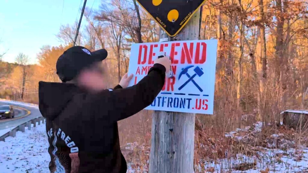A photo of who is alleged to be Garrett Berna, somewhere in New England, during or before March 2025. It is visibly winter, with snow on dying grass, and barren trees in the background. The man alleged to be Berna staples a "Defend American Honor" Patriot Front sign to a utility pole.