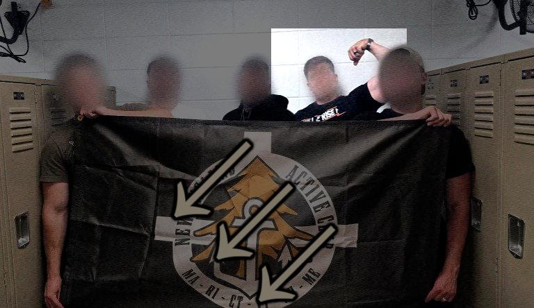 A photo of five men, all white with faces blurred, posing with a New England Active Club flag in a gym, somewhere in Worcester County MA, during June 2025. One man, his head shape largely obvious through the facial blurring, appears to be Garrett Berna. The flag has been defaced with the three arrows.