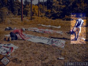 A photo of two white men, incidentally in the exact same outfits from the previous photo taken in Cromwell CT, creating Patriot Front banners in Meriden CT, released during October 2024. One of them, highlighted, wears what appear to be white shorts, a navy blue shirt, and black Adidas trainers. He has dark hair.