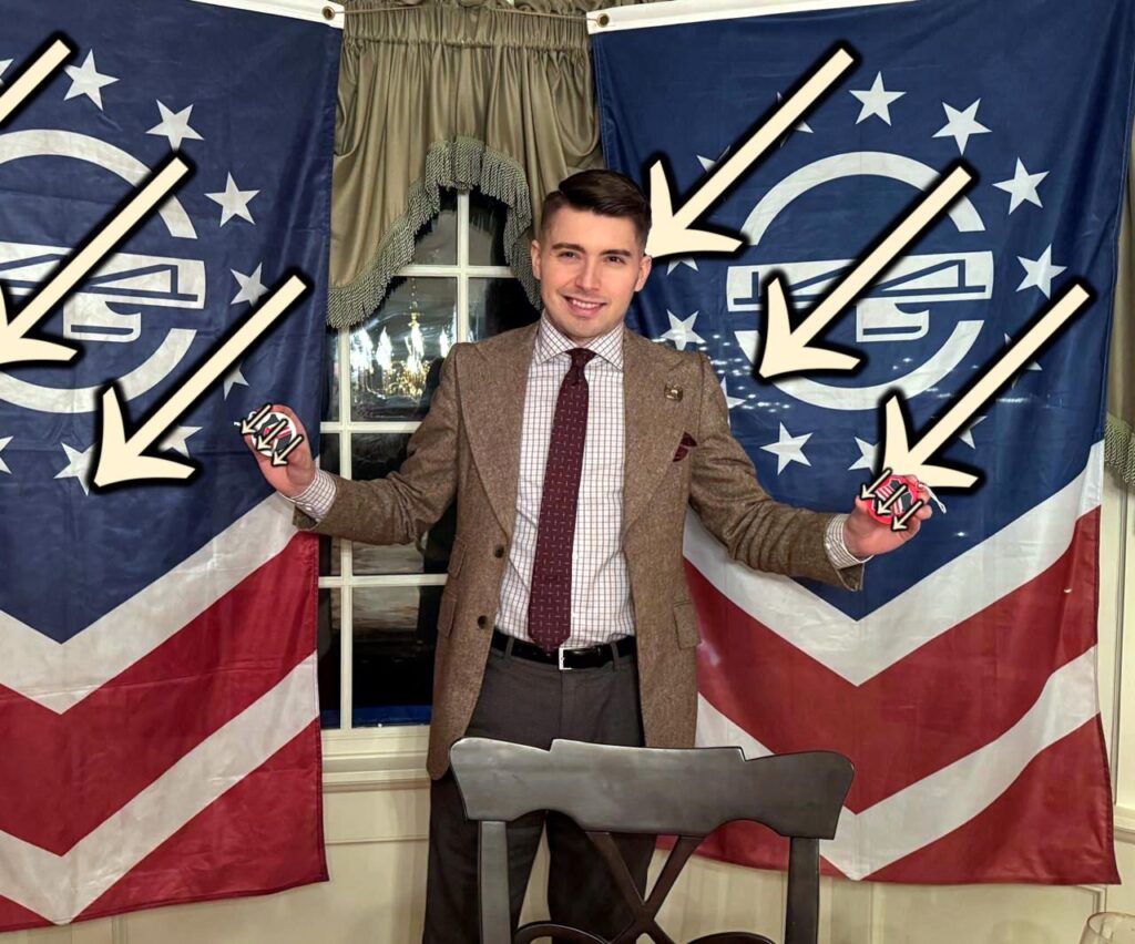 The photo of Jonathan "John Fassbinder" Magnano, which was attached to the previous Telegram post. He is standing between two Patriot Front flags, hung astride a window with a ruffled green valence. He proudly holds what are now apparent as two Christmas ornaments emblazoned with fascist symbols. Said symbols, as well as the fasces of the Patriot Front flags, have been defaced with the three arrows.