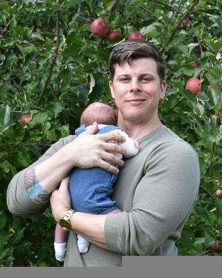 Garrett Berna's current Psychology Today profile photo. He holds his child. A tattoo peeks out of his right sleeve, which is partially rolled up toward his elbow. He has chin. It has a considerable amount of descent, in a way that appears somewhat disproportionate with the rest of his face.