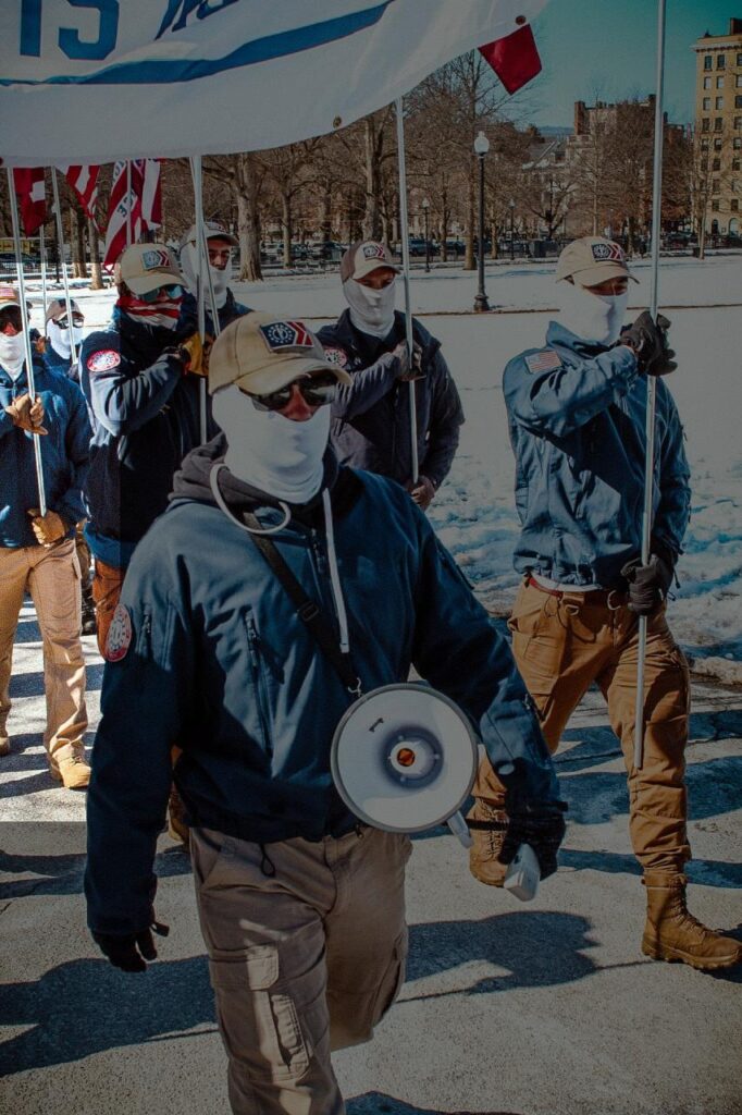 A photo of members of Patriot Front marching through Boston Common during February 2025. They wear the same uniform: khaki cargo pants, blue athletic overshell jackets, white balaclavas or half-face gaiters, and tan baseball caps. Some members vary their uniforms with personal touches, like with fascist hat patches and various kinds of sunglasses. One member in the background appears to be Berna, the shape of his chin remarkably familiar underneath his white balaclava.