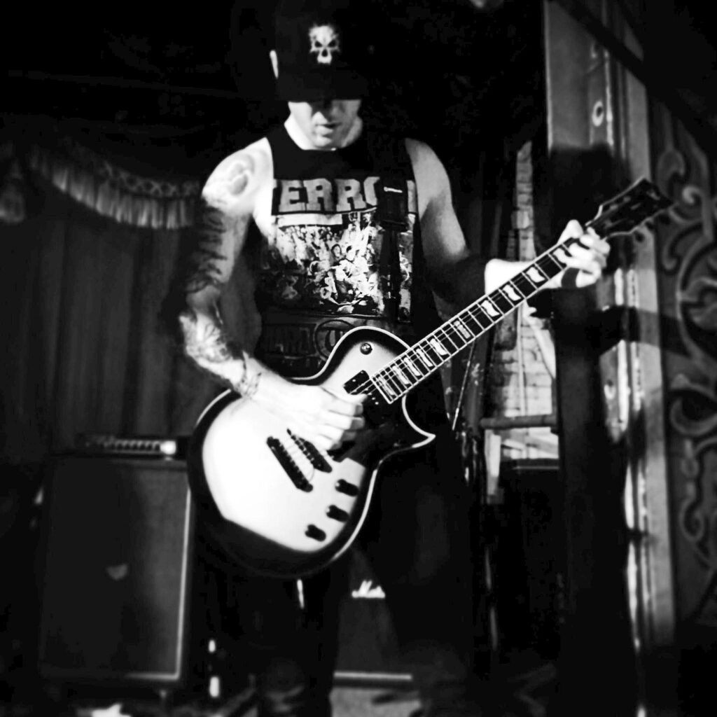 A photo of Chris Rosati uploaded to his social media on July 29th, 2024. Rosati is at what appears to be a concert venue, on stage, playing a guitar. He wears a muscle shirt, dark pants, and a black baseball cap with a skull design on it. Rosati's right arm is visibly tattooed in a 3/4 sleeve ending mid-forearm. His left arm does not have obvious tattoos.